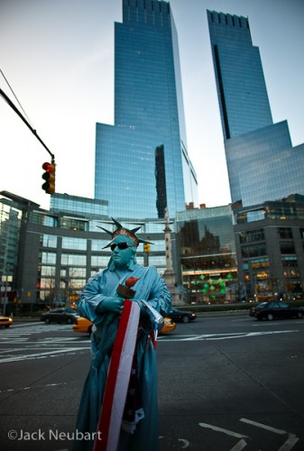 Statue of Liberty in the flesh. We have lots of these living statues stationed around Central Park, here in NYC. I found this one interesting against the backdrop of the towering Time Warner Center. Note the vignetting at the corners, which meant that I obviously shot this at or near maximum aperture (so that the backdrop would blur slightly) with the 21mm lens. Unfortunately, the EXIF data does not record lens aperture, despite the electronic pathways that exist between lens and camera.  ©2009 Jack Neubart. All rights reserved.